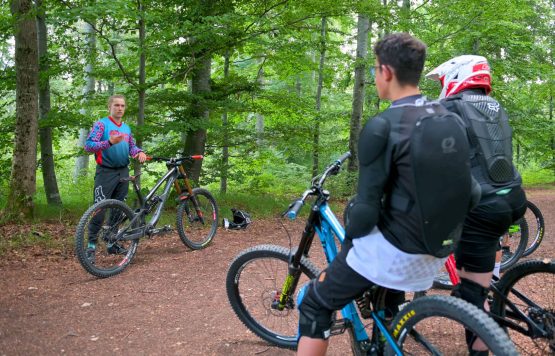Drei Mountainbiker stehen im Wald. Einer gibt Anweisungen, während die anderen auf ihren Fahrrädern warten.