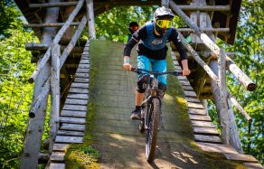Mountainbiker in Schutzkleidung fährt eine Rampe hinunter, umgeben von Holzstruktur und Wald.