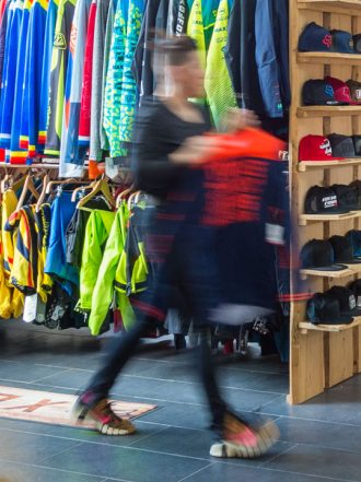 Fahrradgeschäft innen mit bunten Jacken, Helmen und Zubehör auf Regalen; Person hält Kleidungsstück.