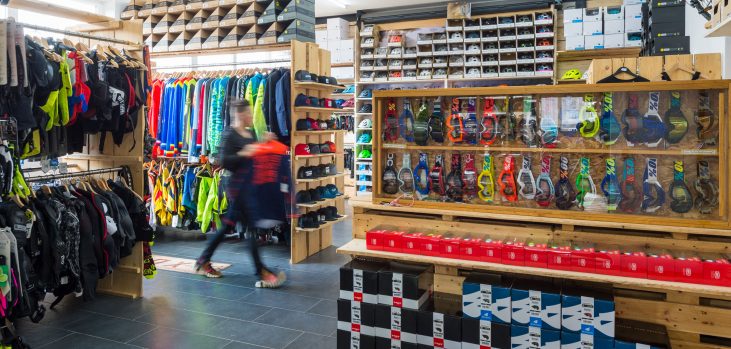 Fahrradgeschäft innen mit bunten Jacken, Helmen und Zubehör auf Regalen; Person hält Kleidungsstück.