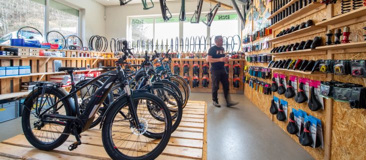 Fahrradladen mit Mountainbikes auf Holzplattform, Zubehör an der Wand, Reifen und Rahmen im Hintergrund. Eine Person steht da.