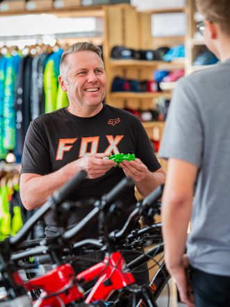 Ein lächelnder Mann zeigt einem Jugendlichen ein grünes Fahrradteil, während sie von Fahrrädern umgeben sind.