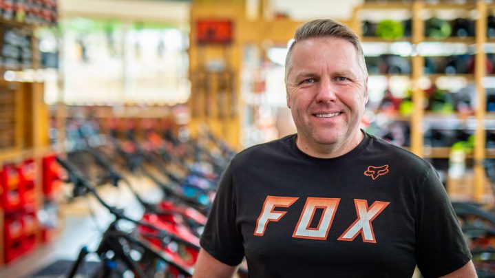 Lächelnder Mann in einem Fahrradgeschäft, umgeben von Fahrrädern und Regalen im Hintergrund.