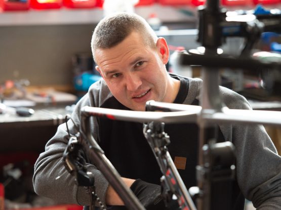 Ein Mann arbeitet an einem Fahrradrahmen in einer Werkstatt, umgeben von Werkzeugen und Zubehör.