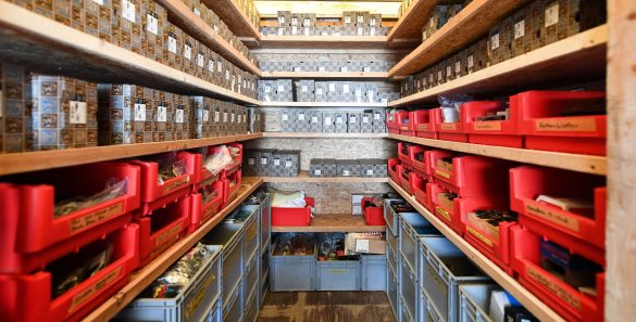 Ein schmaler Raum mit Regalen, die zahlreiche Kisten und Behälter in verschiedenen Farben enthalten, hauptsächlich rot und blau.