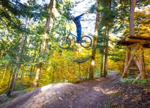 Mountainbiker springt von Rampe in Wald, Arme ausgestreckt, herbstliche Bäume im Hintergrund.