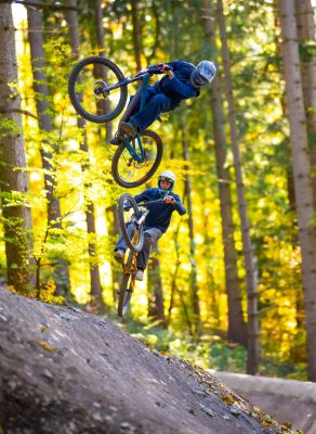 Zwei Mountainbiker springen in einem sonnigen Wald über eine Erhöhung.