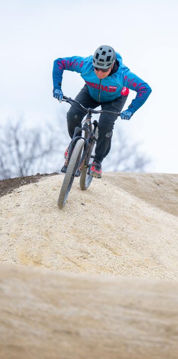 Person in blauer Jacke fährt mit einem Mountainbike eine sandige Hügelstrecke hinunter, Bäume im Hintergrund.