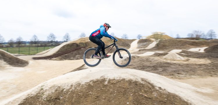Person fährt mit einem Fahrrad über eine hügelige Schotterstrecke in einem weitläufigen Park bei bewölktem Himmel.