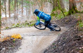 Ein Mountainbiker fährt in einer Kurve auf einem schottrigen Weg durch einen Wald mit Bäumen und trockenem Laub.