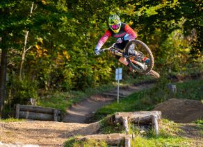 Mountainbiker springt mit Fahrrad über Rampe im Wald, trägt bunte Schutzkleidung und Helm.