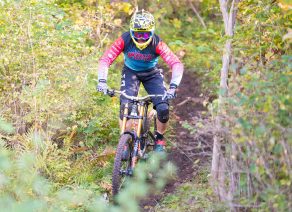Mountainbiker mit Helm und Schutzkleidung fährt energisch durch dichten, grünen Wald entlang eines schmalen Pfades.