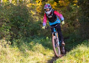 Person in Schutzkleidung fährt Mountainbike auf einem schmalen Waldweg, umgeben von Büschen und Bäumen.