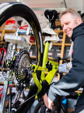 Ein Mechaniker arbeitet an einem Fahrrad, das auf einem Ständer steht. Im Hintergrund sind weitere Fahrräder und Werkzeuge sichtbar.