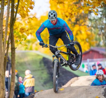 Mountainbiker springt über Erdhügel im herbstlichen Wald, Menschen schauen im Hintergrund zu.