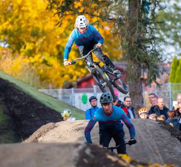 Zwei Mountainbiker springen über Hügel, während Zuschauer aus verschiedenen Positionen zuschauen. Herbstliche Bäume im Hintergrund.