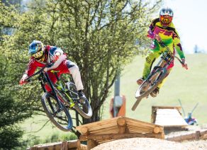 Zwei Mountainbiker springen synchron von einer Holzrampe in einem bewaldeten Gelände.
