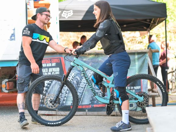 Ein Mann spricht mit einer Frau, die auf einem Mountainbike sitzt. Beide tragen sportliche Kleidung und Schutzkleidung.