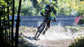 Kind in Schutzausrüstung fährt mit einem Mountainbike rasant durch einen Waldweg mit viel Staub.