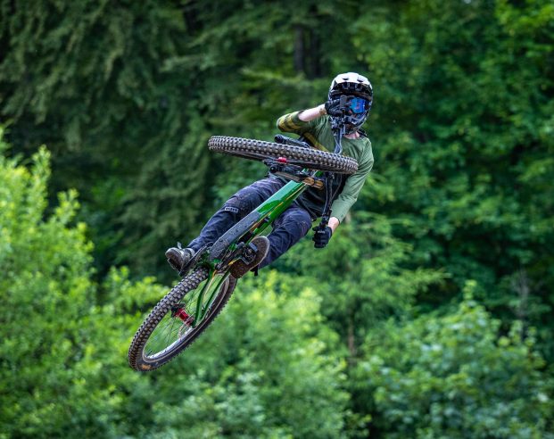 Person macht spektakulären Mountainbike-Sprung im Wald, umgeben von dichtem, grünem Laub.