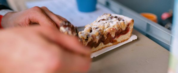 Ein Stück Streuselkuchen mit Zwetschgen auf einem Papptablett, gehalten von einer Hand.