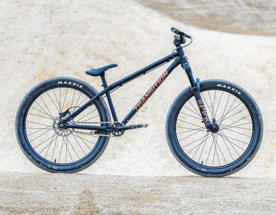 Ein schwarzes Mountainbike mit orangefarbenem Schriftzug steht auf einem sandigen Untergrund.