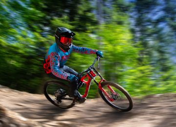 Ein Mountainbiker fährt schnell um eine Kurve, umgeben von grünen Bäumen und einem leichten Schotterweg.